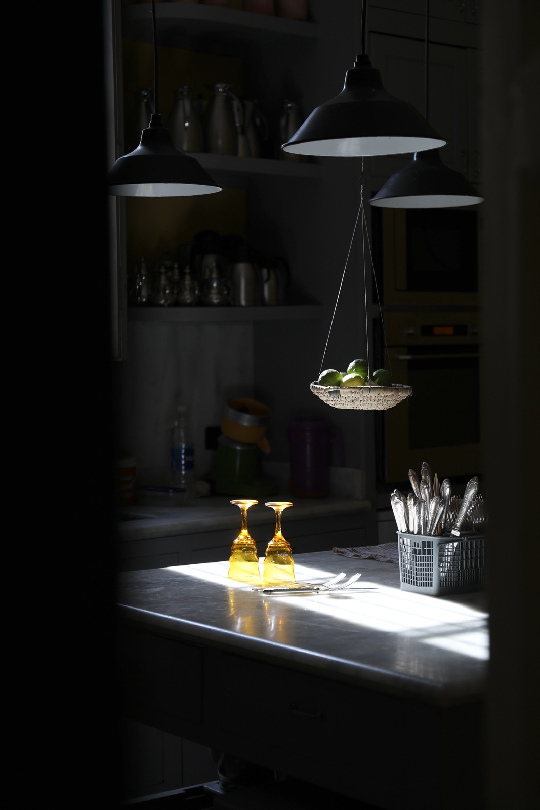 L'office de la maison dans la pénombre — suspensions noires, panier de citrons verts, verres ambrés dans un rai de soleil